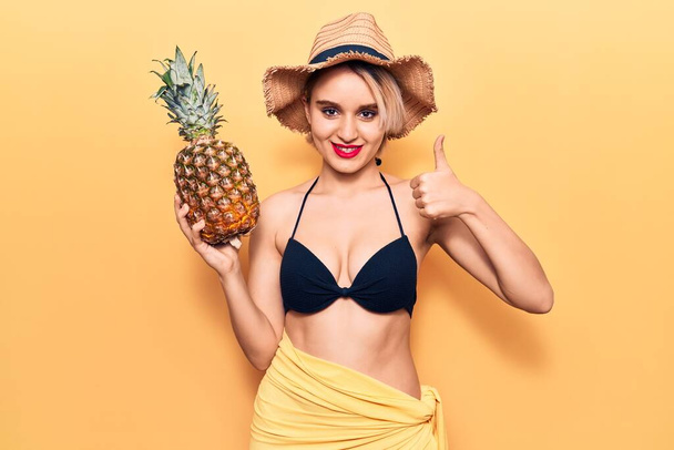 Młoda piękna blondynka nosi bikini i letni kapelusz trzymając ananasa uśmiechnięty szczęśliwy i pozytywny, kciuk do góry robi doskonałe i znak aprobaty  - Zdjęcie, obraz
