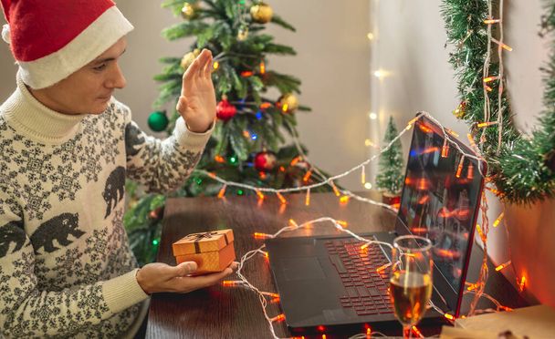 Un jeune homme avec une coupe de champagne et un chapeau rouge de Père Noël est assis devant un écran d'ordinateur portable, souhaitant joyeux Noël à ses amis - Photo, image