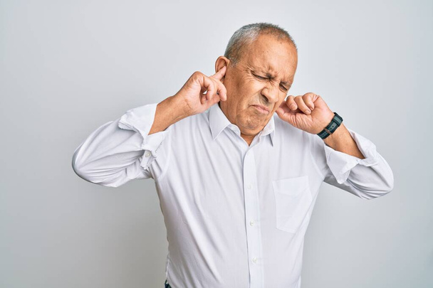 Ein gutaussehender älterer Mann in lässigem weißem Hemd, der die Ohren mit genervten Fingern vor lauter Musik verdeckt. Gehörloses Konzept.  - Foto, Bild