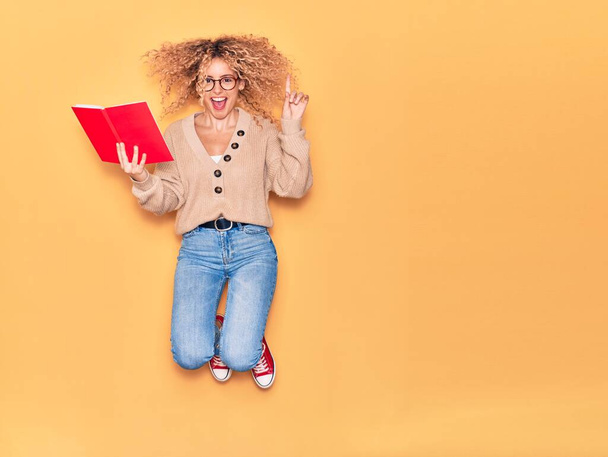 Young beautiful curly student girl wearing glasses smiling happy. Jumping with smile on face reading book over isolated yellow background. - Φωτογραφία, εικόνα