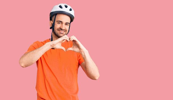 Jonge knappe man draagt een fietshelm glimlachend in liefde met hartsymbool en vorm met handen. romantisch concept.  - Foto, afbeelding