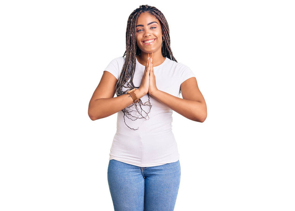 Jovem afro-americana com tranças vestindo roupas casuais orando com as mãos juntas pedindo perdão sorrindo confiante.  - Foto, Imagem