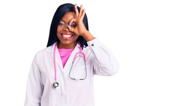 Młoda Afroamerykanka ubrana w stetoskop robi ok gest z uśmiechem dłoni, oko patrząc przez palce ze szczęśliwą twarzą.  - Zdjęcie, obraz