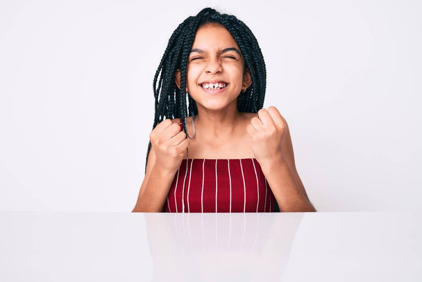 Jong Afrikaans Amerikaans meisje kind met vlechten dragen casual kleren zitten op de tafel enthousiast voor succes met opgeheven armen en ogen gesloten vieren overwinning glimlachen. winnaarsconcept.  - Foto, afbeelding