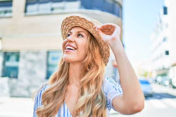 Young beautiful blonde woman on vacation wearing summer hat smiling happy. Standing with smile on face at street of city. - Foto, Imagen