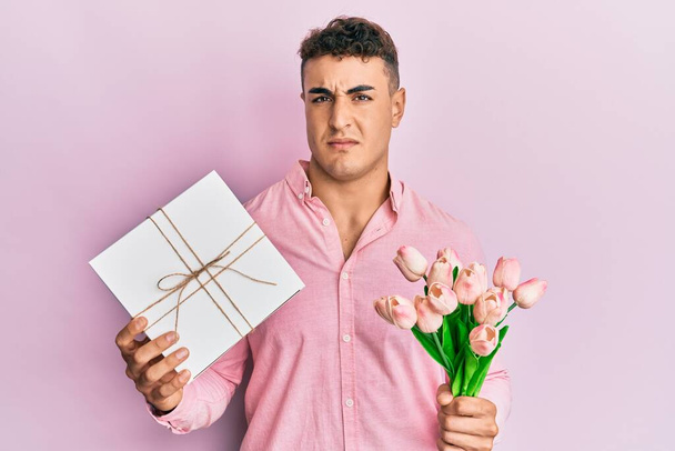 Spaanse jongeman met jubileum cadeau en boeket bloemen clueless en verwarde uitdrukking. twijfelconcept.  - Foto, afbeelding