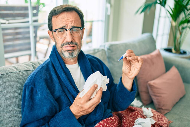 Hombre de mediana edad sintiéndose enfermo de resfriado y fiebre en casa, enfermo de gripe sentado en el sofá - Foto, imagen