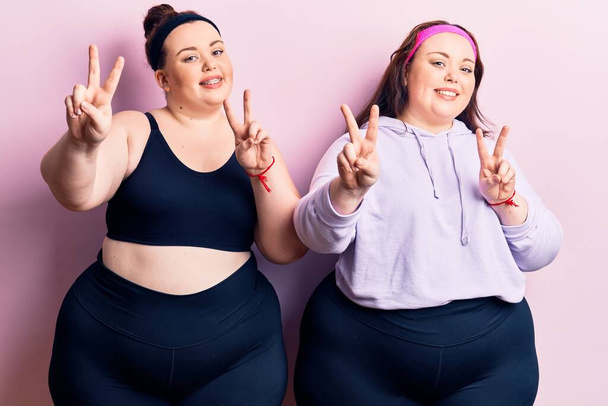 Young plus size twins wearing sportswear smiling looking to the camera showing fingers doing victory sign. number two.  - Photo, Image