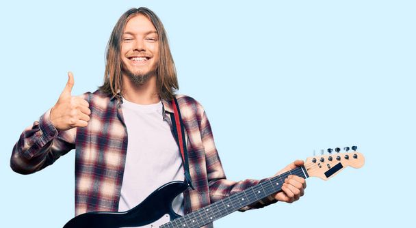Uzun saçlı yakışıklı beyaz adam elektronik gitar çalıyor mutlu ve pozitif gülümsüyor, başparmağıyla mükemmel ve onay işareti yapıyor.  - Fotoğraf, Görsel