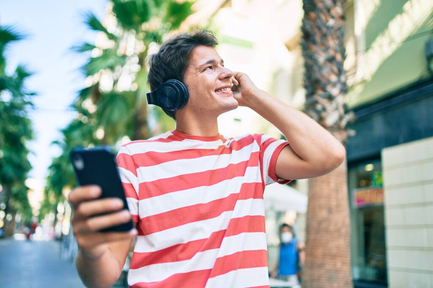 Junger Kaukasier lächelt glücklich mit Smartphone und Kopfhörer in die Stadt. - Foto, Bild