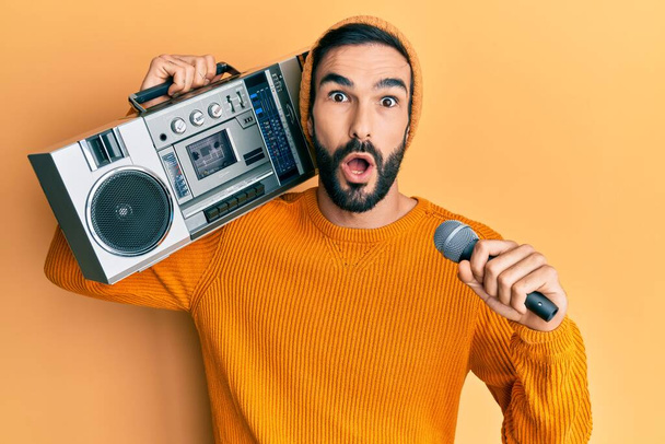 Young hispanic man holding boombox, listening to music singing with microphone afraid and shocked with surprise and amazed expression, fear and excited face.  - Photo, Image