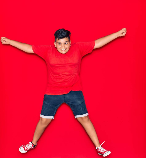 Adorable kid wearing casual clothes smiling happy with arms opened. Jumping with smile on face over isolated red background - Фото, изображение
