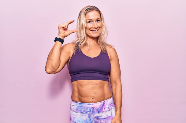Μέση ηλικία καυκάσιος ξανθιά γυναίκα φορώντας αθλητικά ρούχα πάνω από ροζ φόντο χαμογελώντας και αυτοπεποίθηση gesturing με το χέρι κάνει μικρό σημάδι μέγεθος με τα δάχτυλα αναζητούν και η κάμερα. έννοια μέτρου.  - Φωτογραφία, εικόνα