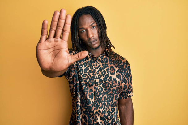 Hombre afroamericano con trenzas con camisa estampada de leopardo haciendo parar de cantar con la palma de la mano. expresión de advertencia con gesto negativo y grave en la cara.  - Foto, imagen