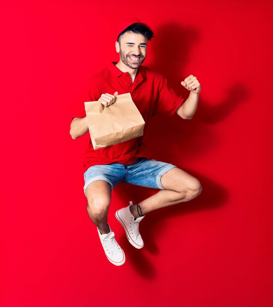 Young handsome deliveryman wearing cap smiling happy. Jumping with smile on face holding paper bag with food over isolated red background - Zdjęcie, obraz