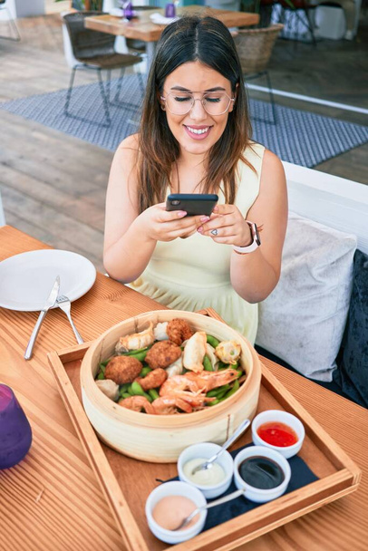 Jeune belle femme hispanique souriant heureux. Assis sur la table en utilisant un smartphone pour faire des photos de nourriture au restaurant - Photo, image