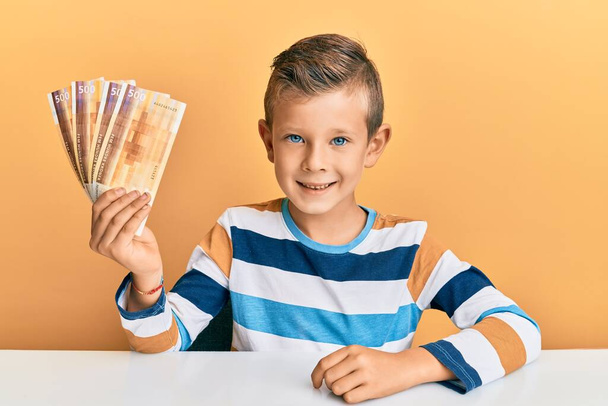 Urocze dziecko rasy kaukaskiej trzymające 500 norweskich banknotów koronowych siedzących na stole, wyglądające pozytywnie i szczęśliwie, stojące i uśmiechnięte z ufnym uśmiechem pokazującym zęby  - Zdjęcie, obraz