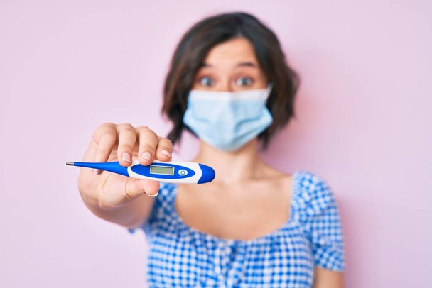 Young beautiful woman wearing medical mask holding holding thermometer scared and amazed with open mouth for surprise, disbelief face  - Photo, Image