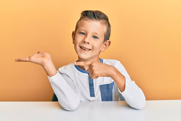 Adorable caucasian kid wearing casual clothes sitting on the table amazed and smiling to the camera while presenting with hand and pointing with finger.  - Foto, Imagem
