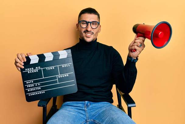 Handsome man with tattoos holding video film clapboard and megaphone clueless and confused expression. doubt concept.  - Foto, afbeelding
