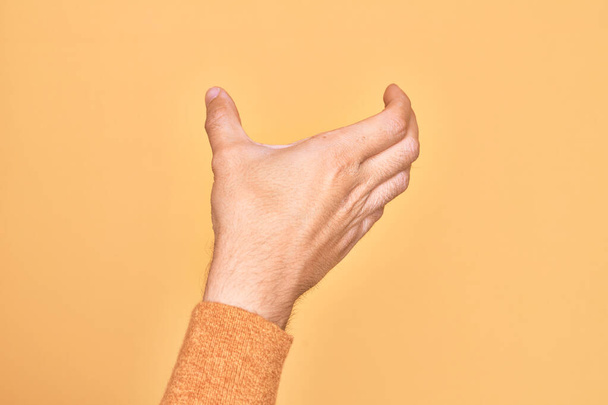 Рука кавказского юноши показывает пальцы на изолированном желтом фоне, держа невидимый предмет, пустая рука делает обрезание и хватающий жест - Фото, изображение