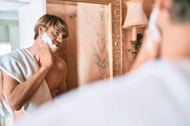 Όμορφος ξανθός άντρας κοιτάζει τον καθρέφτη στο μπάνιο εξοικονομώντας αφρό και ξυράφι. - Φωτογραφία, εικόνα