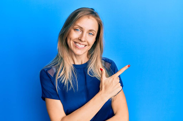 Schöne kaukasische Frau in lässigem blauem T-Shirt lächelt fröhlich und zeigt mit der Hand und dem Finger zur Seite  - Foto, Bild