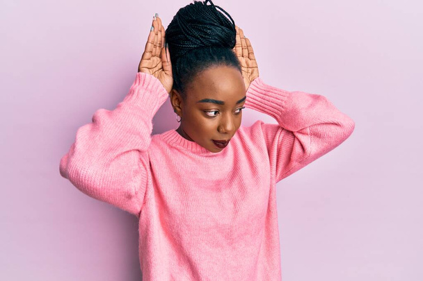 Молода афроамериканська жінка, одягнена в повсякденний зимовий светр, робить жест кролячих вух з пальмами, що виглядають цинічно і скептично. Великодній кролик..  - Фото, зображення