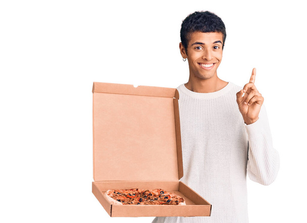 Junger afrikanisch-amerikanischer Mann mit Pizzaschachtel überrascht mit einer Idee oder Frage, Zeigefinger mit glücklichem Gesicht, Nummer eins  - Foto, Bild