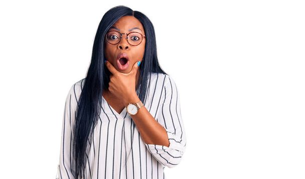 Young african american woman wearing casual clothes and glasses looking fascinated with disbelief, surprise and amazed expression with hands on chin  - Photo, Image