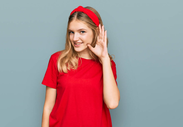 Красивая юная кавказская девушка в непринужденной красной футболке, улыбающаяся рукой над ухом, слушая слухи или сплетни. концепция глухоты.  - Фото, изображение