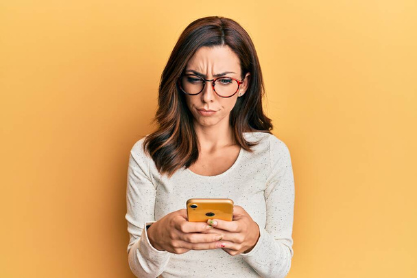 Giovane donna bruna utilizzando smartphone su sfondo giallo scettico e nervoso, accigliato sconvolto a causa di problemi. persona negativa.  - Foto, immagini