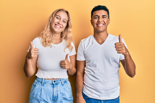 若い異人種間のカップルは、手で積極的なジェスチャーを行うカジュアルな白いTシャツの成功のサインを着て、親指を笑顔と幸せ。陽気な表情と勝者のジェスチャー.  - 写真・画像
