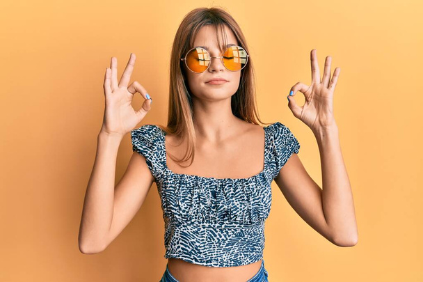 Adolescente chica caucásica con gafas de sol de moda amarillo relajarse y sonreír con los ojos cerrados haciendo gesto de meditación con los dedos. concepto de yoga.  - Foto, imagen