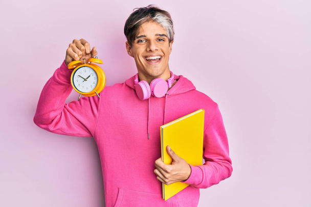Young hispanic man holding book and alarm clock smiling with a happy and cool smile on face. showing teeth.  - Zdjęcie, obraz
