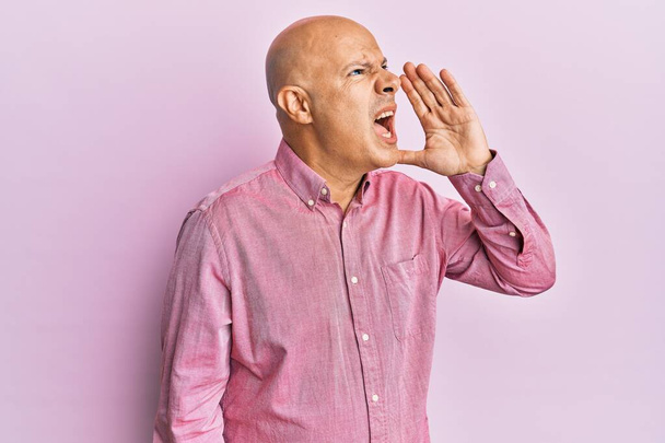 Middle age bald man wearing pink business shirt shouting and screaming loud to side with hand on mouth. communication concept.  - Foto, Imagen