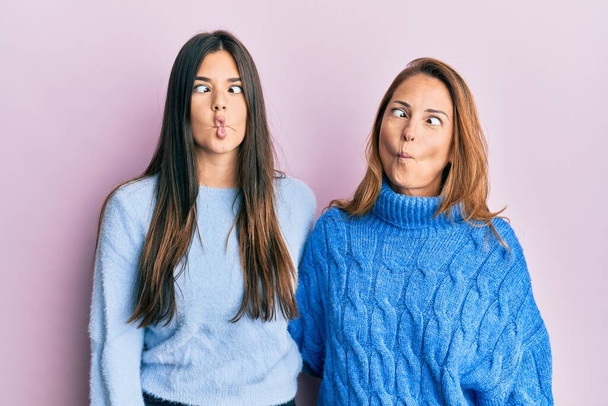 Ισπανόφωνη οικογένεια της μητέρας και της κόρης φορώντας μάλλινο χειμωνιάτικο πουλόβερ κάνοντας ψαρομούρη με χείλη, τρελή και κωμική χειρονομία. αστεία έκφραση.  - Φωτογραφία, εικόνα
