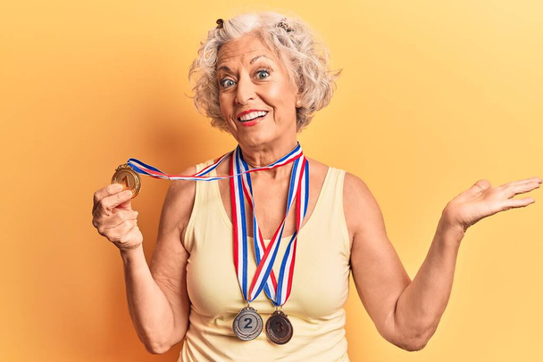 Mulher de cabelos grisalhos sênior vestindo medalhas celebrando a realização com sorriso feliz e expressão vencedora com mão levantada  - Foto, Imagem