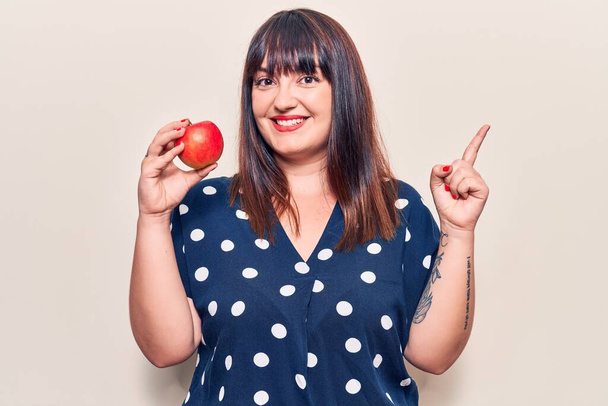 Jovem plus size mulher segurando maçã vermelha sorrindo feliz apontando com a mão e o dedo para o lado  - Foto, Imagem