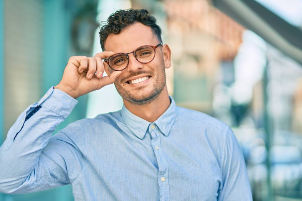 Jovem empresário hispânico sorrindo feliz tocando seus óculos na cidade. - Foto, Imagem