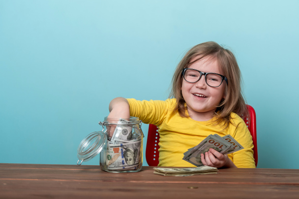 Happy child takes money from glass jar full of dollars. Portrait of kid with money in hand. Savings for future. Financial education for kids. How to think and investing and do not afraid - Photo, Image