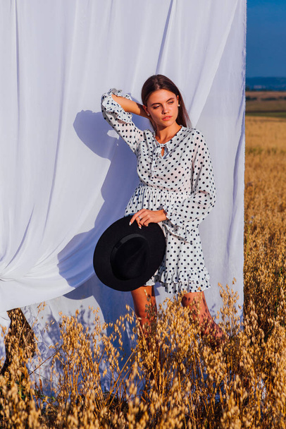 Rural Countryside Scene. Young beautiful woman with long hair dressed in white Polka-dot dress and black hat standing at golden oat field with a white curtain on the background. - Photo, Image
