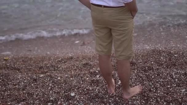 a barefoot man in shorts stands on a pebble beach near the water - Πλάνα, βίντεο