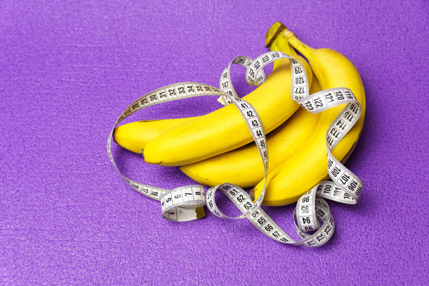 Un ramo de plátanos y una cinta métrica sobre un fondo púrpura. Estilo de vida saludable y nutrición adecuada. - Foto, imagen