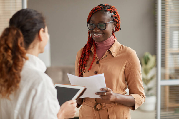 Pas se portrét mladé afroamerické podnikatelky mluví s manažerkou a vesele se usmívá, zatímco stojí v moderním kancelářském interiéru - Fotografie, Obrázek
