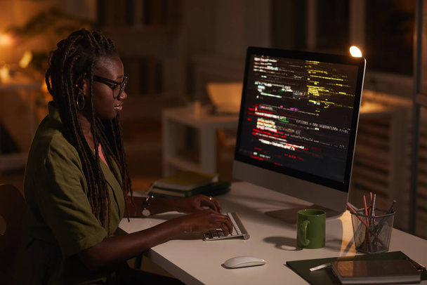 Portrait latéral d'une femme afro-américaine contemporaine écrivant du code et regardant un écran d'ordinateur tout en travaillant dans un bureau sombre, copiez l'espace - Photo, image