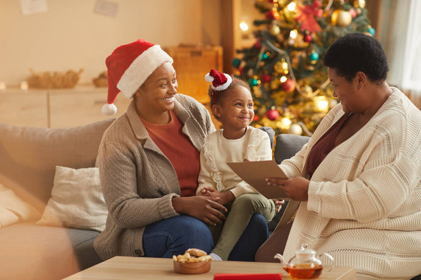 Retrato cálido de una familia afroamericana sonriente leyendo una carta a Santa con una linda niña mientras disfruta de la temporada de Navidad en casa - Foto, imagen