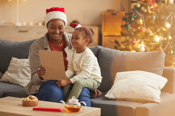 Портрет з теплими тонами усміхненої афро-американської матері, що читає листа Санті з милою маленькою дівчинкою, насолоджуючись різдвяним сезоном вдома, копіюйте простір. - Фото, зображення