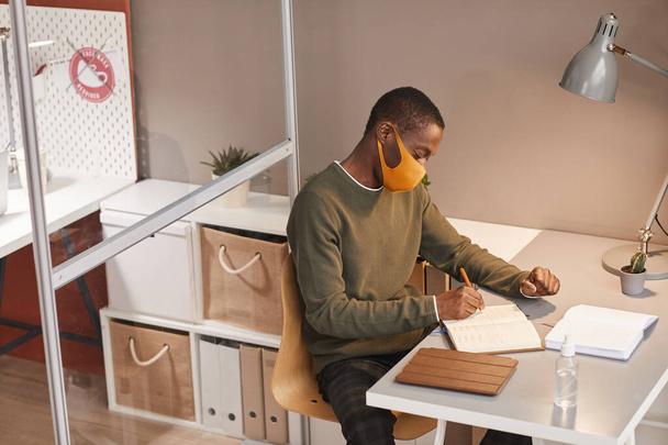 Duży kąt portret młodego Afroamerykanina w masce i piśmie w planerze podczas pracy przy biurku w biurze, przestrzeń do kopiowania - Zdjęcie, obraz