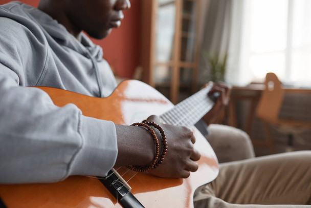 Зблизька молодий афроамериканець грає на акустичній гітарі, сидячи на дивані і створюючи музику вдома, копіюйте простір. - Фото, зображення